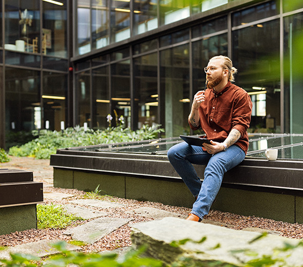 En mand sidder med en tablet i en frodig gård og kigger ud i luften