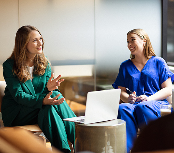 To mennesker sidder og taler sammen foran en computer, hvoraf den ene smiler, mens den anden forklarer noget